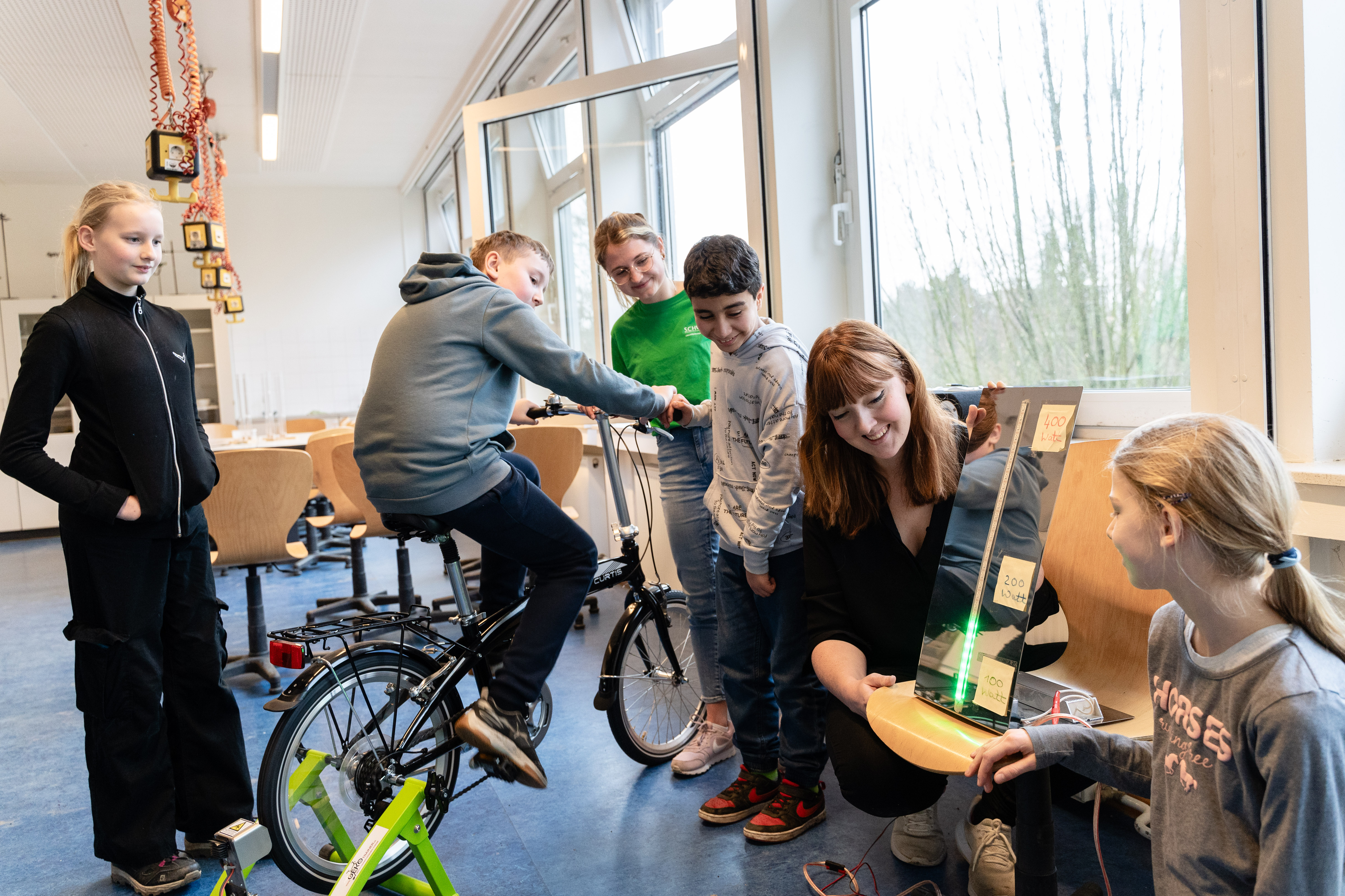 Ein Schüler sitzt auf einem Fahrrad, weitere Schüler*innen und Rebecca Baumer (SCHUBZ) sowie Lisa Persiel (Lünestrom) schauen auf einer Anzeige, wieviel Leistung erradelt wird.